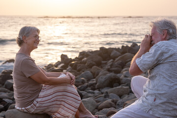 Fototapeta premium Senior man takes a picture to his wife sitting on the beach at sunset light