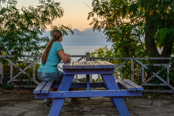 Naklejka na ściany i meble A girl is sitting at a table in the park and admiring the sea with mountains at sunset.