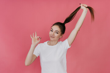 Cheerful Italian young woman in white t-shirt making ok sign , looking at camera, pulling ponytail...