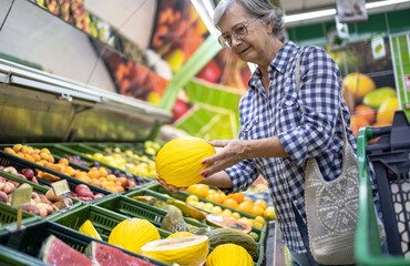 Elderly woman in the supermarket choosing fresh fruit, holding a yellow melon in her hand. Seasonal fruit baskets on background
