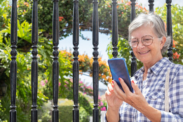 Naklejka na ściany i meble Smiling caucasian senior woman outdoors in a flowered garden using mobile phone