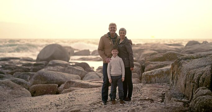 Happy grandparents with kid by the beach, with family and posing for a picture with a smile. Senior man and woman with boy by sea coast or sand or rocks on summer vacation or holiday at sunrise.