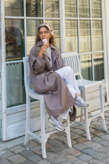 Fototapeta premium african american woman in stylish trench coat eating ice cream and sitting on chair in prague.
