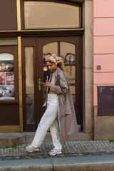 full length of stylish african american woman in headscarf and trench coat walking and using smartphone in prague.