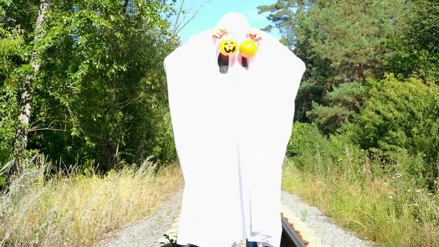 Funny Halloween. A ittle kid in a white suit holding baskets in the shape of a pumpkin, a scary Halloween ghost, on the street. A child dressed as Halloween ghosts scares and asks for candy