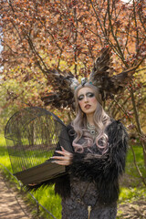 A girl in a gloomy image with a beautiful make-up, a crown of feathers and a cage in her hands