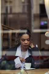 african american woman in trendy blazer stirring coffee in cup while sitting behind window glass in...