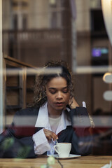 african american woman in trendy blazer stirring coffee in cup while sitting behind window glass in cafe.