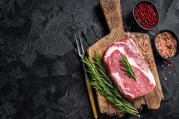 Raw Rib-eye Steak, beef marbled meat on wooden board with rosemary. Black background. Top view....