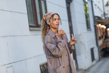 young african american woman in stylish trench coat and headscarf applying lip gloss on street in prague.