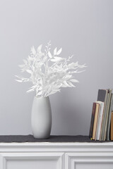 Cropped close-up shot of a white branch of willow in a white vase. The vase with a decorative willow branch stands on a shelf with books on a gray background. Front view.