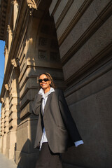 sunshine on face of happy african american woman in sunglasses and oversize suit walking on urban street in prague.