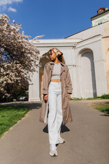 full length of stylish african american woman in trench coat standing in park near blooming tree.