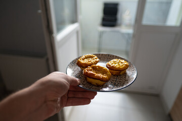 A hand holds a plate with fresh cupcakes on it. In the background is a balcony