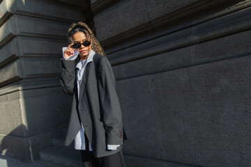 young african american woman in oversize suit adjusting sunglasses near building on urban street in prague.