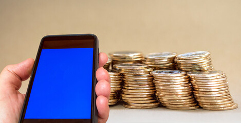 Cell phone with blue screen in hand and background with stacks of coins.