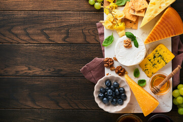 Assortment of cheese, honey, cracker, blueberries, grapes with red and white wine in glasses antipasto server on white marble board on old rustic wooden table background. Flat lay, copy space.