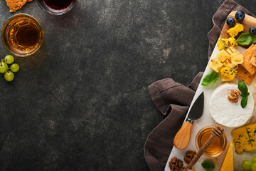 Assortment of cheese, honey, cracker, blueberries, grapes with red and white wine in glasses antipasto server on white marble board on old dark grey background. Flat lay, copy space.