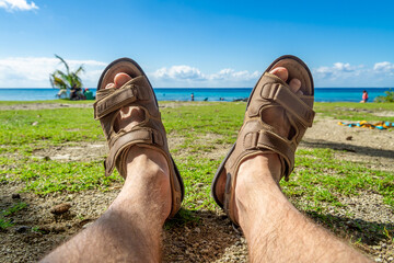 Men's feet in sandals against the background of the sea and cloudy sky. A man sunbathes on the shore overgrown with grass. The concept of recreation and tourism.