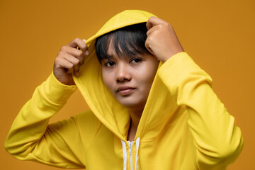 Portrait of a handsome Asian tomboy dressed in casual hoodie looking at camera on yellow studio...