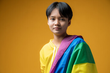 Portrait of a handsome young tomboy with rainbow flag on shoulder looking at camera against yellow background.