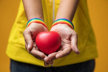 close-up woman LGBT lesbian hand wearing rainbow-patterned wristband and red heart in hand on...