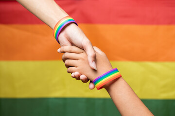 Two Asian woman holding hands with rainbow-patterned wristband on their wrists with LGBTQ rainbow flag in the background. lgbt and homosexuality concept