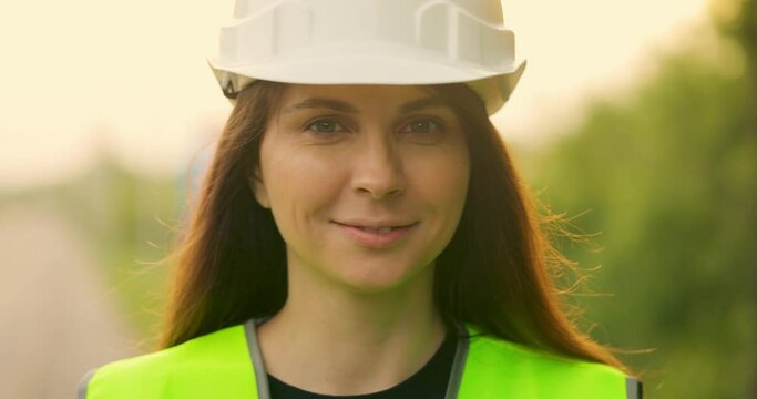 Young beautiful girl in helmet and light green vest looking at camera and smiles. Woman worker takes off her helmet. Close-up. Slow motion