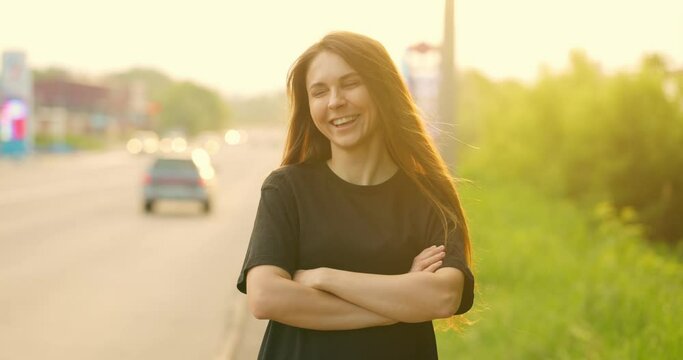 Confident charming attractive woman with her arms crossed on her chest in casual clothes poses for camera. Successful smiling girl looks at camera outdoor. Slow motion