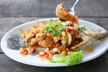 Spoon to scoop up a piece of fried fish from plate of Thai food name Deep-Fried snapper with Three-Flavoured Sauce. Deep Fried Fish with Chili Sauce served in a white plate on a blurred background. 
