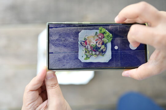 Woman hand holding smartphone or mobile phone to  take pictures the spicy seafood glass noodle salad with  and share in social network before eating. Spicy seafood vermicelli salad on a wooden table.
