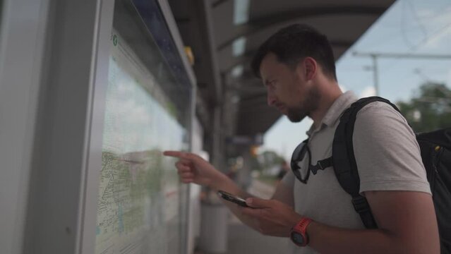Male passenger analyzes transport card at a public transportation stop and matches it to an app on his phone in Munich, Germany. Checking map and waiting for transport in city. Public transport route