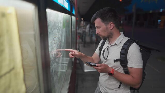 A Caucasian man looks at a transport card at a bus stop at night in the City of Munich, Germany. The topic is public transport in Bavaria. Traveler with backpack at the urban transport map