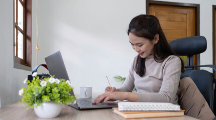 Beautiful woman writing/taking notes while sitting in front her computer laptop at the wooden working table over living room bookshelf as background.