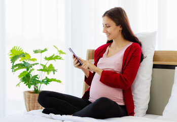 A beautiful pregnant caucasian woman is happily using her smartphone in bed while resting. - 527318823