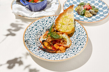Seafood platter with cooked shrimps, mussels, sea scallops in tomato sauce on white table. Stew seafood in italian style with ciabatta bread. Seafood dish: prawns, shellfish, scallops on ceramic plate