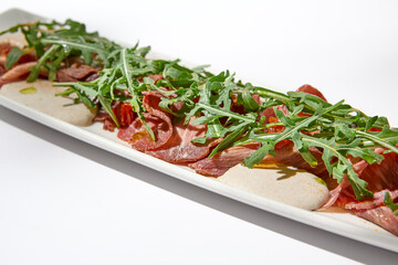 Sliced veal meat on white table with harsh shadows. Sliced veal meat with tuna sauce and arugula. Vitello tonnato on ceramic plate in summer day. Aesthetic food menu
