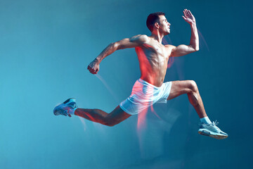 Full length portrait side view of fitness guy running. Half naked young man in white shorts on blue background.