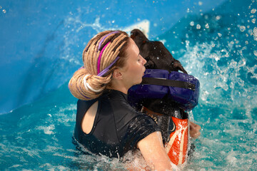 Dog in life jacket swim in the swimming pool with coach. Pet rehabilitation. Recovery training...