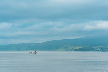Naklejka premium waterscape of the sea bay with a fishing ship and a mount in the clouds in the background, a view of the Mendeleev volcano from the side of the town of Yuzhno-Kurilsk