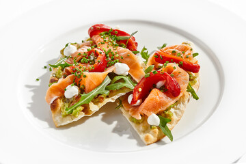 Bruschetta with salmon, avocado and paprika on white plate. Salmon bruschetta on crispy focaccia. Appetizer with guacamole, trout, cheese and paprika on toast. Fish antipasti in restaurant menu.