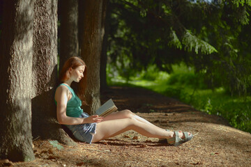 Side view of white redhead young woman in green top and blue shorts reading in the forest.