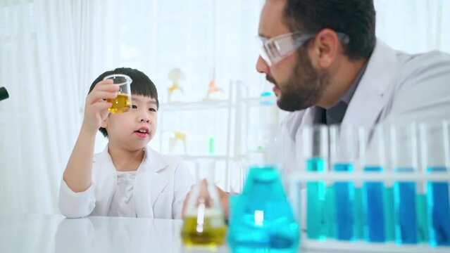 Young Asia kid doing experiment with foreigner teacher in the laboratory at school. Smart boy mixing colorful liquid in the flask and get green color. Chemical and learning elementary class