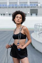 Portrait of african american sportswoman holding jump rope and looking at camera on stadium.