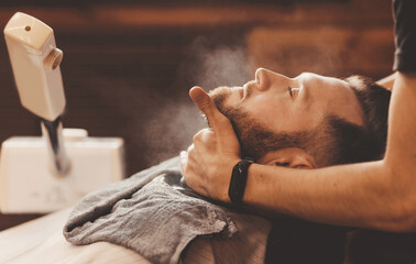 Barber hairdresser steams skin of man face before dangerous shave with razor. Barbershop concept vintage toning