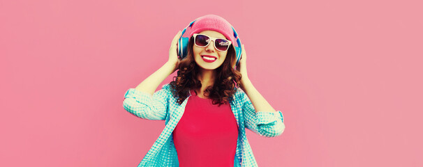 Colorful portrait of happy smiling young woman listening to music in headphones wearing casual hat on pink background