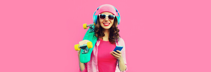 Summer colorful portrait of happy smiling young woman listening to music in headphones with smartphone and skateboard on pink background, blank copy space for advertising text