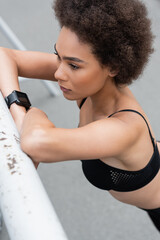 high angle view of curly african american woman in fitness tracker leaning on fence outdoors.