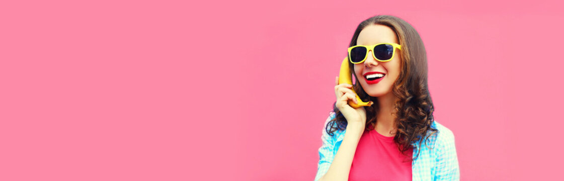 Colorful portrait of funny young woman calling on banana phone looking away on pink background, blank copy space for advertising text