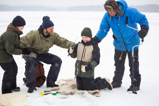 Happy mature men looking at boy fishing while kneeling on frozen lake during winter